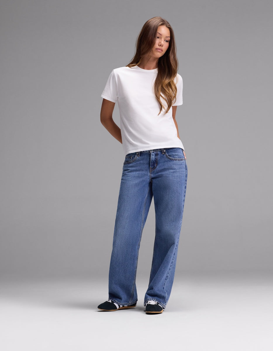 A person stands against a plain gray background, wearing the Cozy Earth Women’s Go-To Tee in white, blue wide-leg jeans, and black slip-on shoes with white accents.