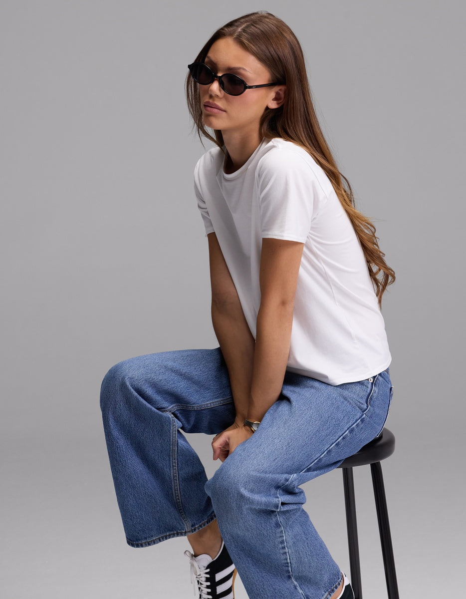 A person with long brown hair wears sunglasses, blue jeans, and black sneakers, sitting on a black stool against a plain gray background. She is wearing the Cozy Earth Women’s Go-To Tee.