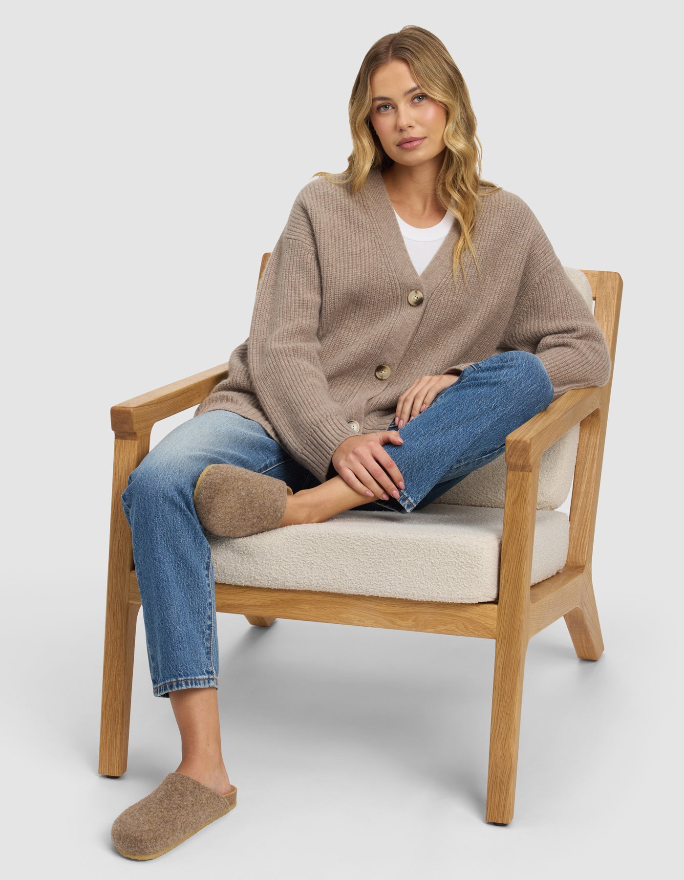 A woman with long blonde hair sits relaxed on a wooden armchair, wearing blue jeans and Cozy Earth’s Lakehouse Clog slippers. One leg is crossed over the other, and she looks calmly at the camera.