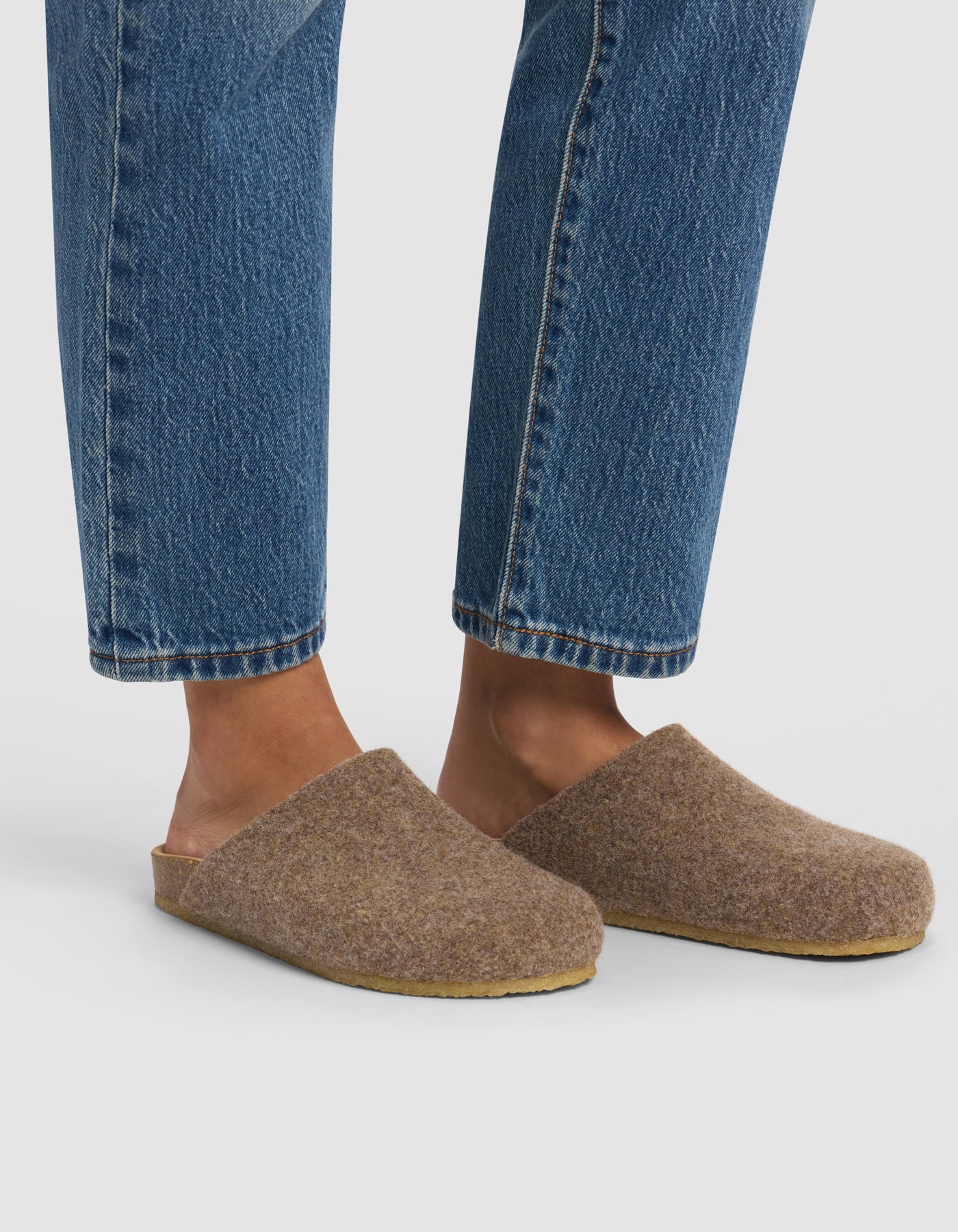 A person wearing blue jeans and Cozy Earth Lakehouse Clog slippers stands on a white background, with only their lower legs and feet visible.