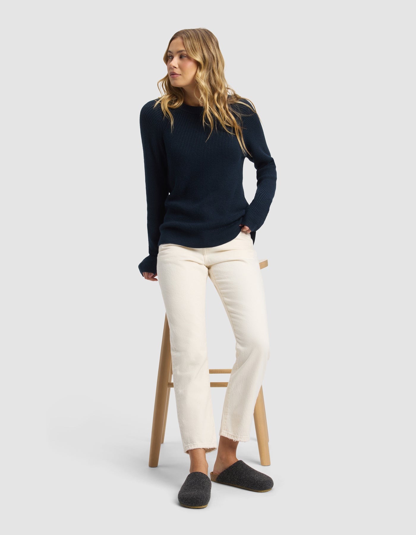 A woman with wavy blonde hair sits on a wooden stool, wearing a navy blue knit sweater, off-white pants, and Cozy Earth's Lakehouse Clog slippers in dark gray, set against a plain light gray background.