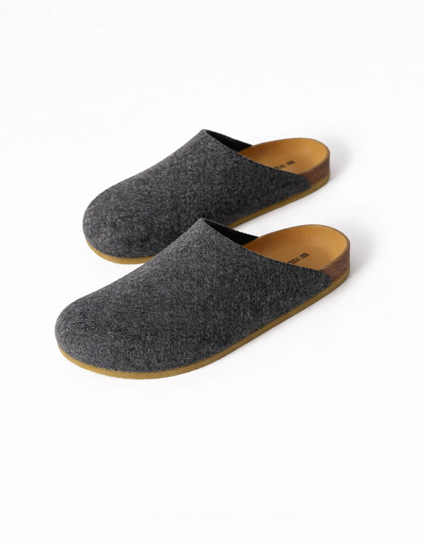 The Lakehouse Clog by Cozy Earth features a minimalist design with dark grey woolen uppers and natural brown soles. It has a closed toe, open back, and is ideal for indoor wear. A logo is visible on the insole, all showcased on a white background. 