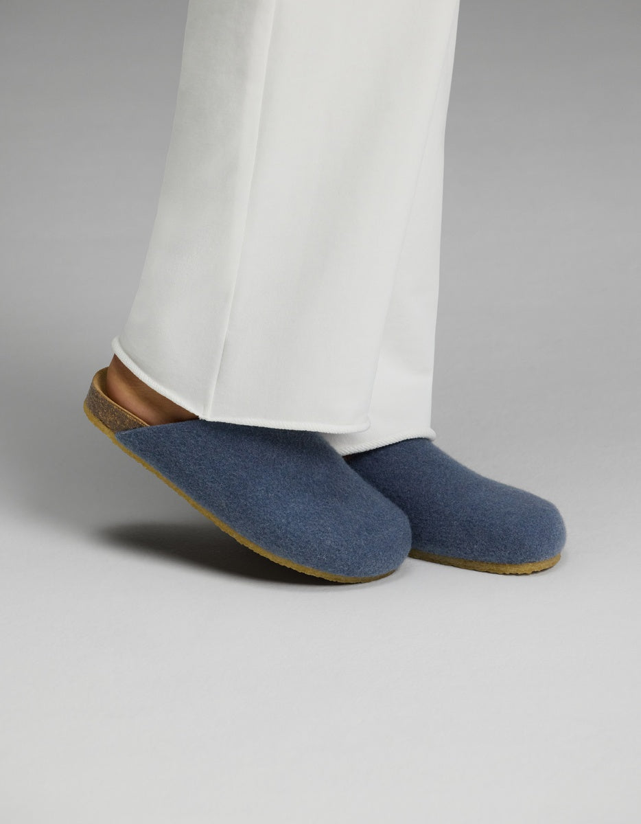 A person wearing white pants and Cozy Earth Lakehouse Clog slippers in light blue, featuring cork soles, stands on a light gray surface.|Color:Light Blue