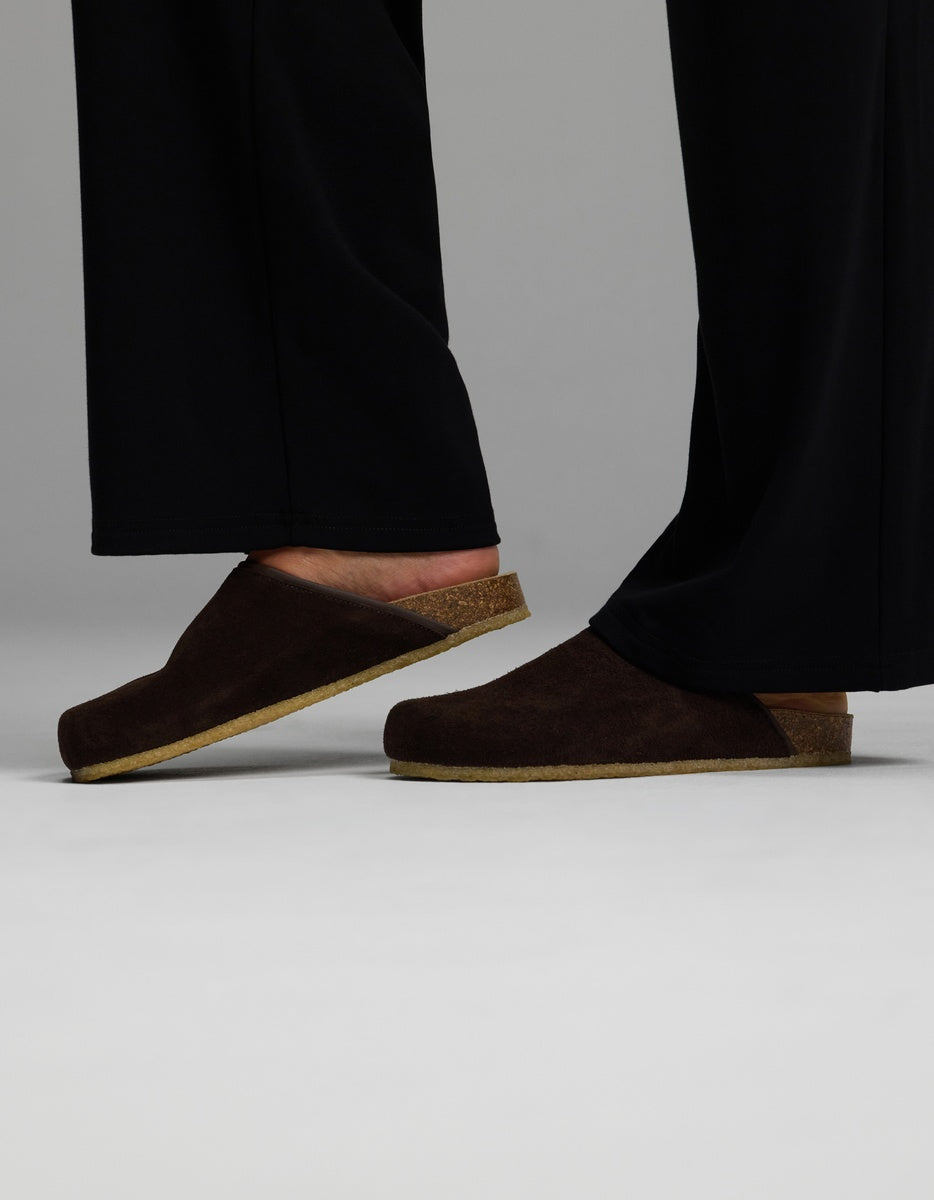 A person wearing black pants and the Cozy Earth Lakehouse Clog in Suede—dark brown slip-on clogs with cork soles—stands on a light, neutral background, with only their lower legs and feet visible.|Color:Espresso