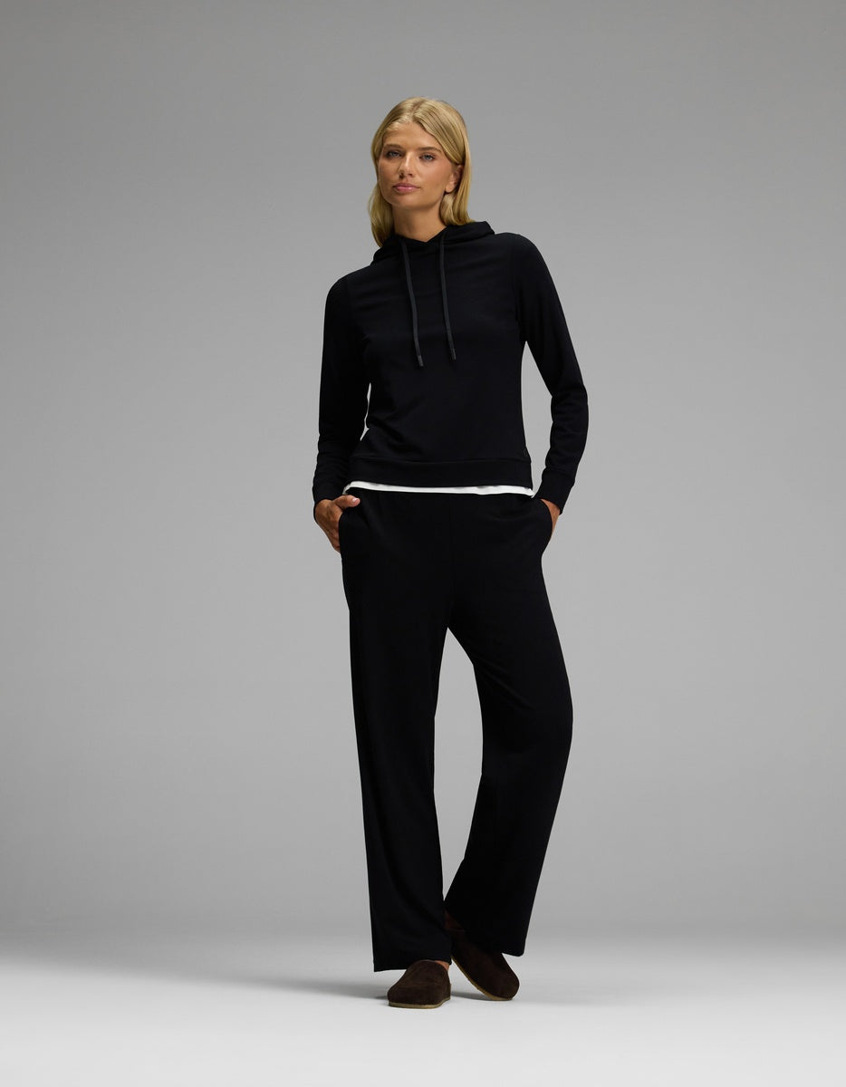 A woman stands with hands in her pockets, wearing the Lakehouse Clog in Suede by Cozy Earth, paired with a black long-sleeve hoodie and matching black pants layered over a white shirt, against a plain gray background.|Color:Espresso