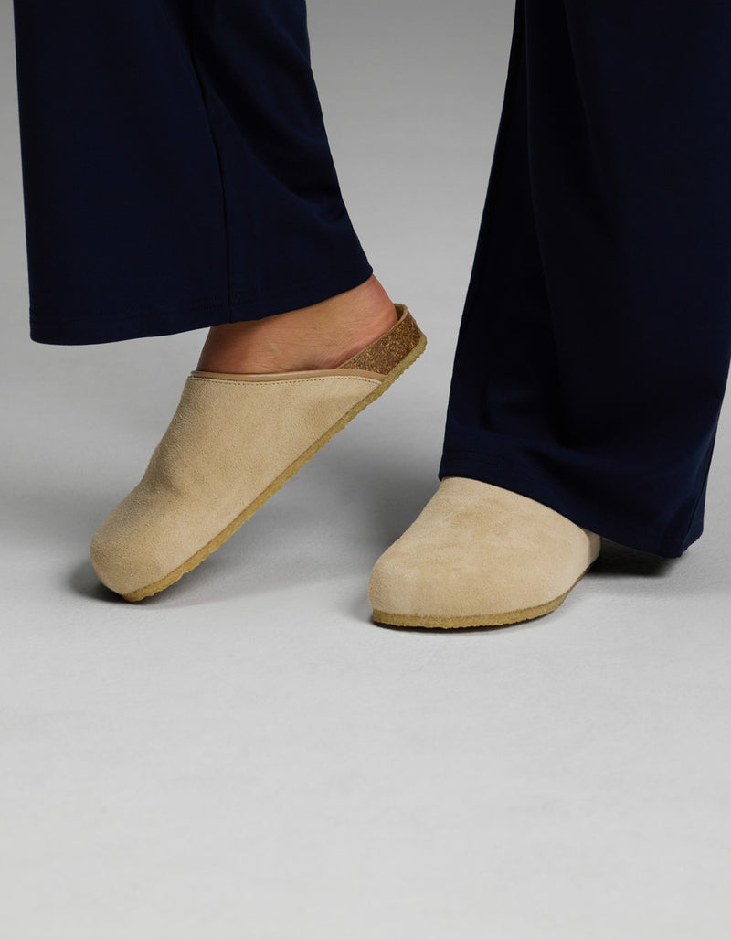 A person wearing navy blue pants and Cozy Earth’s Lakehouse Clog in Suede stands on a light gray surface.