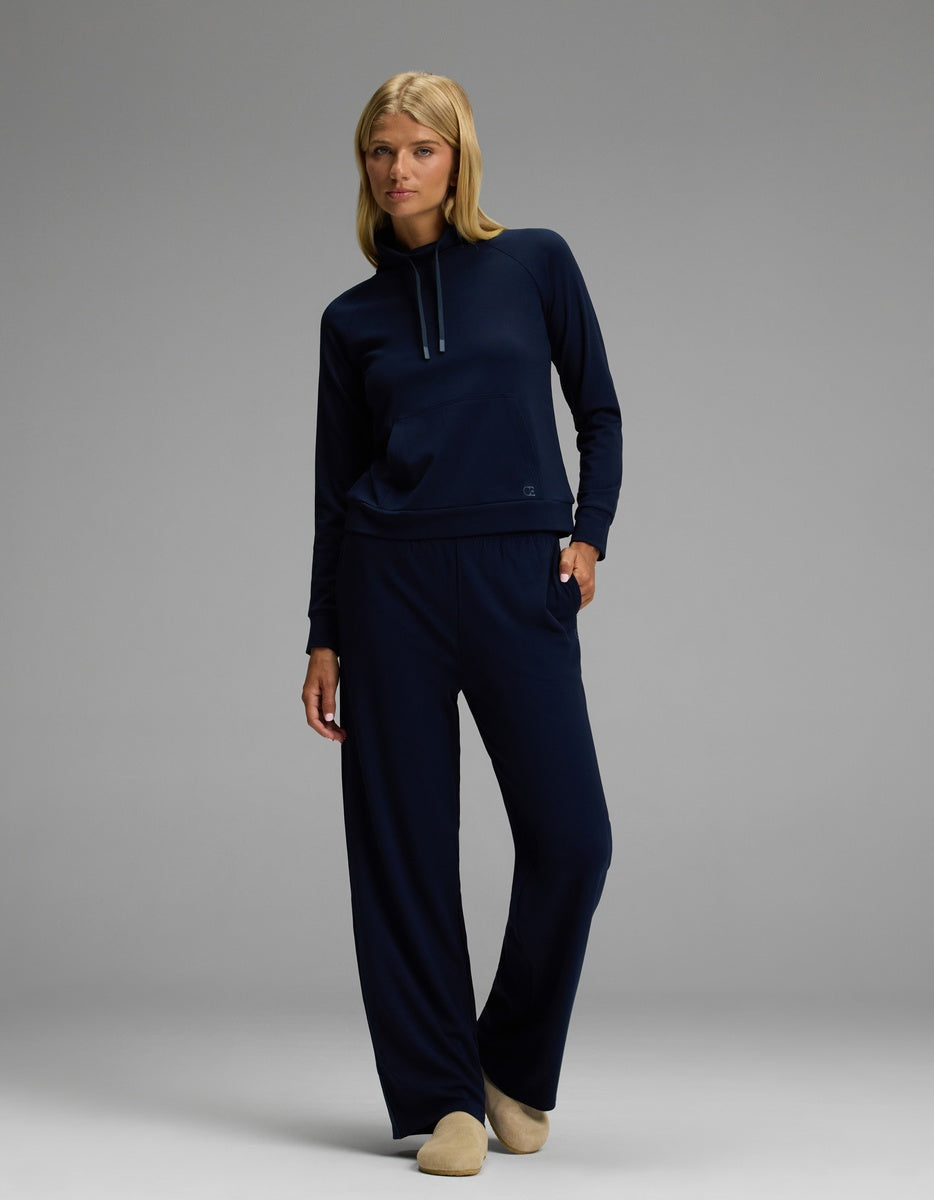 A woman with blonde hair and glasses stands in a relaxed pose against a gray background, dressed in a navy blue hoodie and matching wide-leg pants. She completes her look with Cozy Earth’s Lakehouse Clog in Suede.