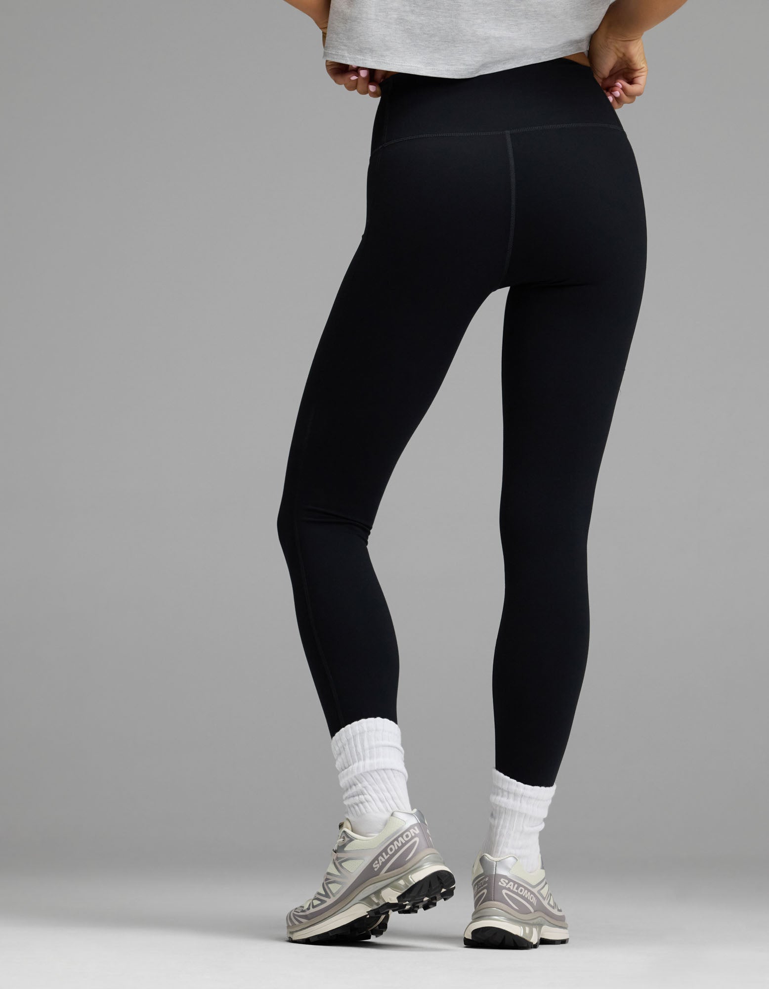 A person stands with their back to the camera wearing Cozy Earth’s Women’s Liv Legging, paired with white socks, gray sneakers, and a light gray cropped shirt, set against a plain gray background.|Color:Jet Black