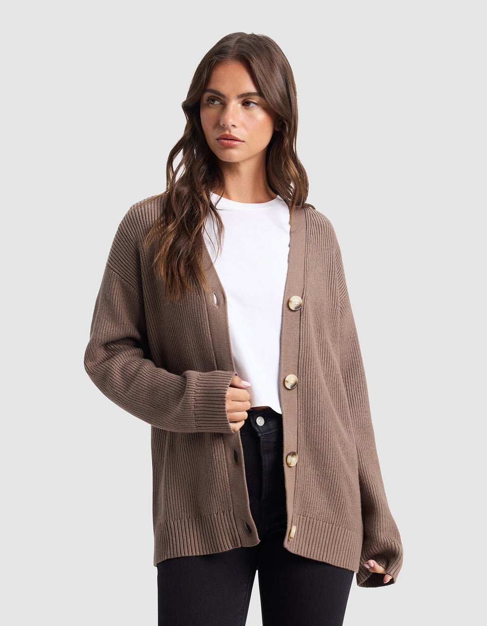 A woman stands against a light gray background, wearing Cozy Earth’s Oversized Classic Cardigan in brown with large buttons, layered over a white t-shirt and black pants.