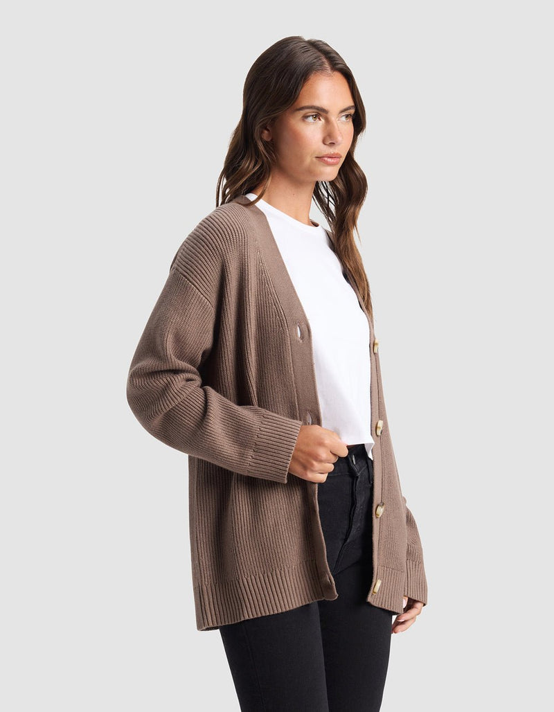 A woman stands against a light gray background, wearing the Cozy Earth Oversized Classic Cardigan in taupe over a white t-shirt and black pants, looking slightly to the side.