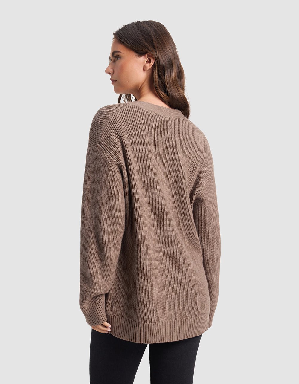 A woman with long brown hair is wearing Cozy Earth's Oversized Classic Cardigan in brown, paired with black pants, and is standing with her back to the camera against a plain light gray background.