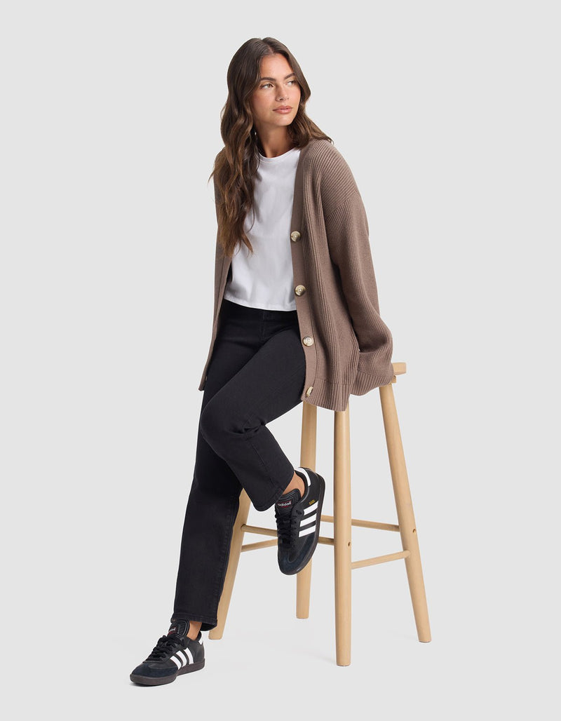 A woman with long brown hair, wearing Cozy Earth's Oversized Classic Cardigan in taupe, a white t-shirt, black jeans, and black Adidas sneakers, sits on a wooden stool against a plain light gray background.