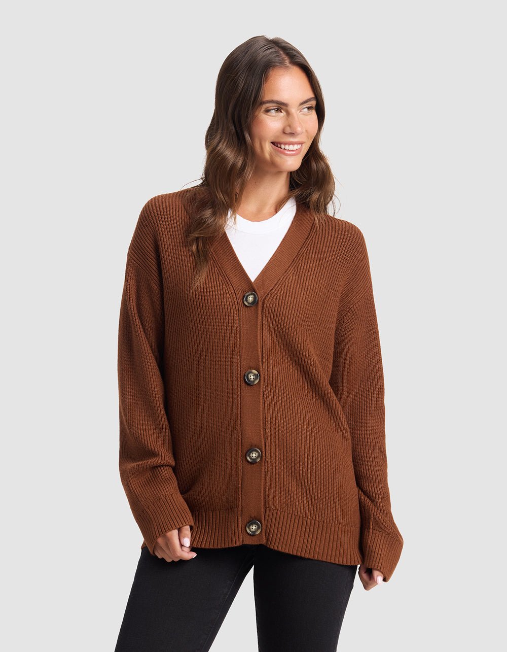 A woman smiles against a plain light gray background, wearing Cozy Earth's Oversized Classic Cardigan in brown over a white shirt and black pants.