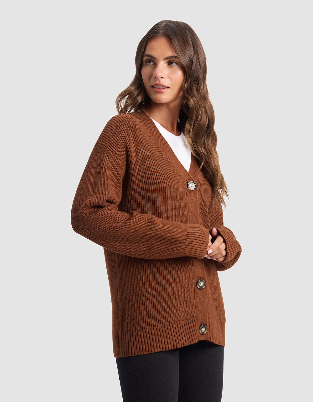 A woman with long brown hair wears the Cozy Earth Oversized Classic Cardigan, featuring a ribbed texture and large buttons, layered over a white top and black pants against a plain light gray background.
