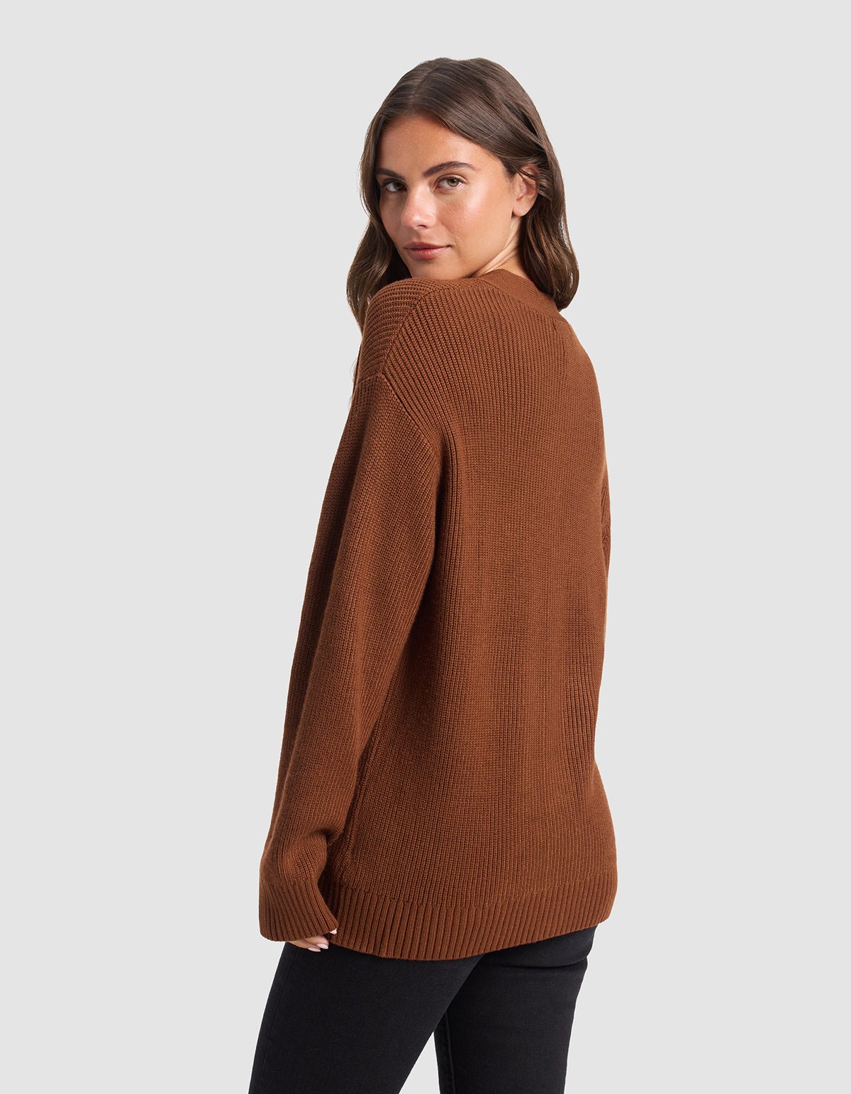 A woman with long brown hair models Cozy Earth's Oversized Classic Cardigan, a loose-fitting, brown ribbed sweater, paired with black pants. She stands sideways and looks over her shoulder against a plain light gray background.