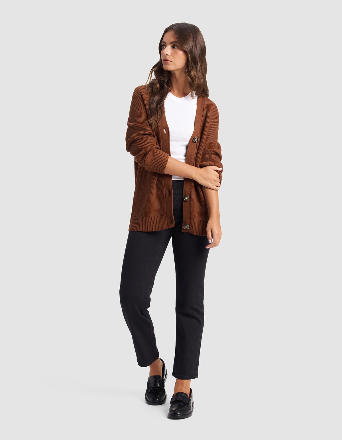 A woman stands against a plain background wearing the Cozy Earth Oversized Classic Cardigan over a white tee, black pants, and black loafers. She has long hair, looks slightly to the side, and folds one arm.