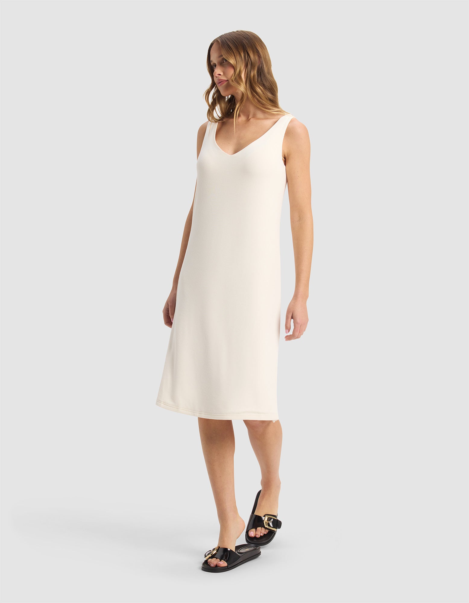 A woman in the Cozy Earth Women’s Bamboo Rib-Knit V-Neck Tank Lounge Dress stands against a plain light gray background, smiling and looking to the side.