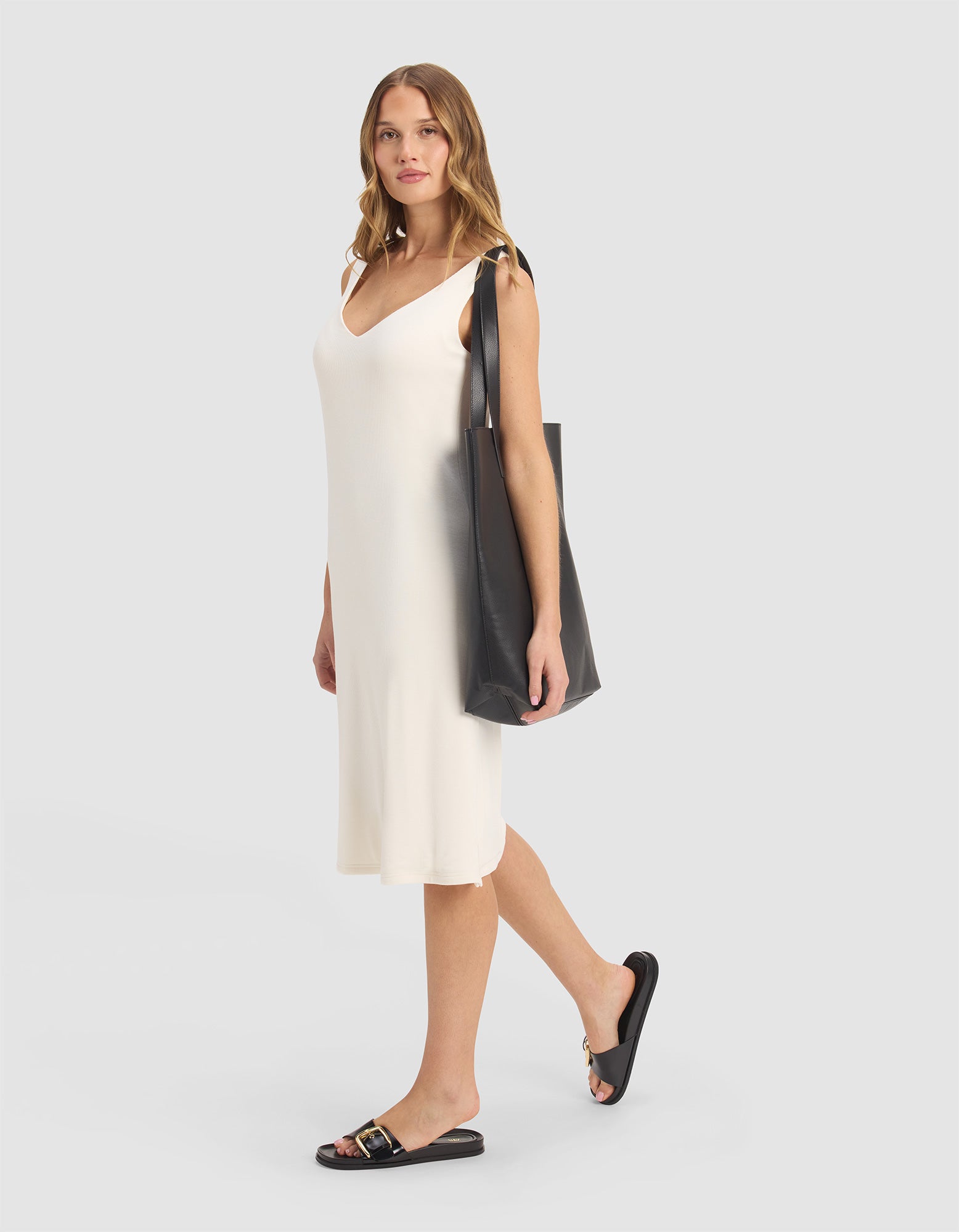 A woman wears the Cozy Earth Women’s Bamboo Rib-Knit V-Neck Tank Lounge Dress in cream and black slide sandals, carrying a large black tote bag on her shoulder against a plain light gray background.
