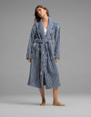 A person stands barefoot against a plain gray background, wearing the Cozy Earth Ribbed Terry Bath Robe—a long, blue-and-white striped robe with a belt—over casual clothes.|Color:Obsidian Stripe