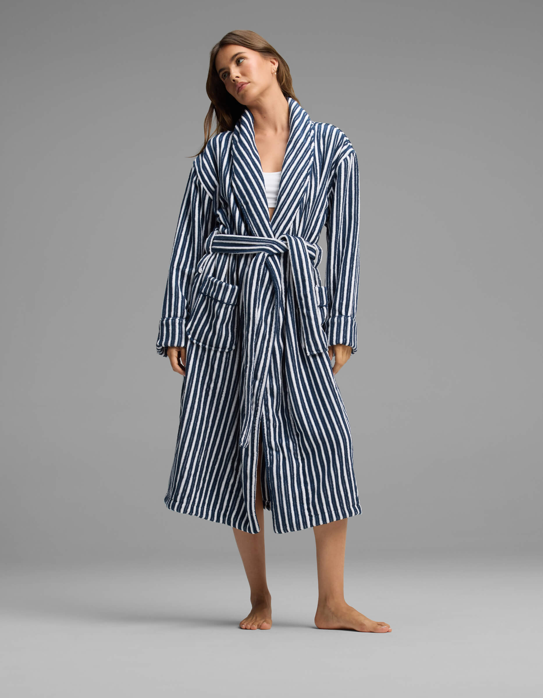 A person stands barefoot against a plain gray background, wearing the Cozy Earth Ribbed Terry Bath Robe—a long, blue-and-white striped robe with a belt—over casual clothes.