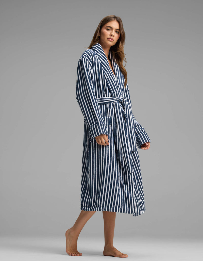 A woman stands barefoot wearing the Cozy Earth Ribbed Terry Bath Robe, featuring a long, blue and white striped design with a belt, against a plain gray background.