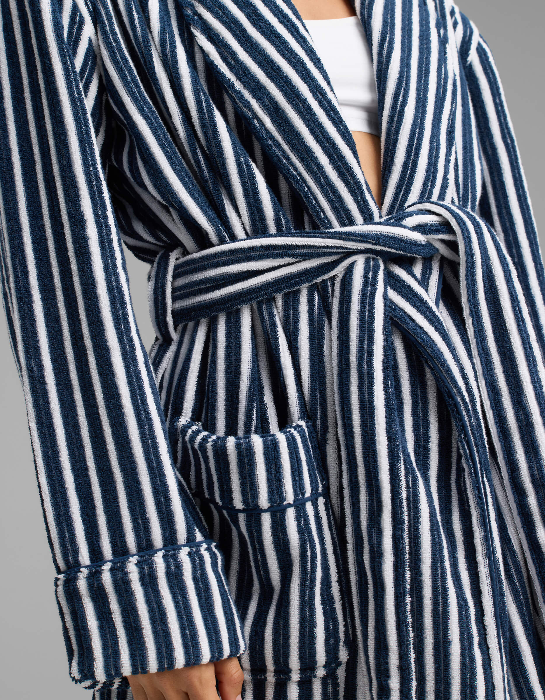A person wearing the Cozy Earth Ribbed Terry Bath Robe, featuring blue and white vertical stripes, a waist tie belt, and a visible front pocket. The robe is layered over a white top, with only the torso and part of the arm shown.|Color:Obsidian Stripe