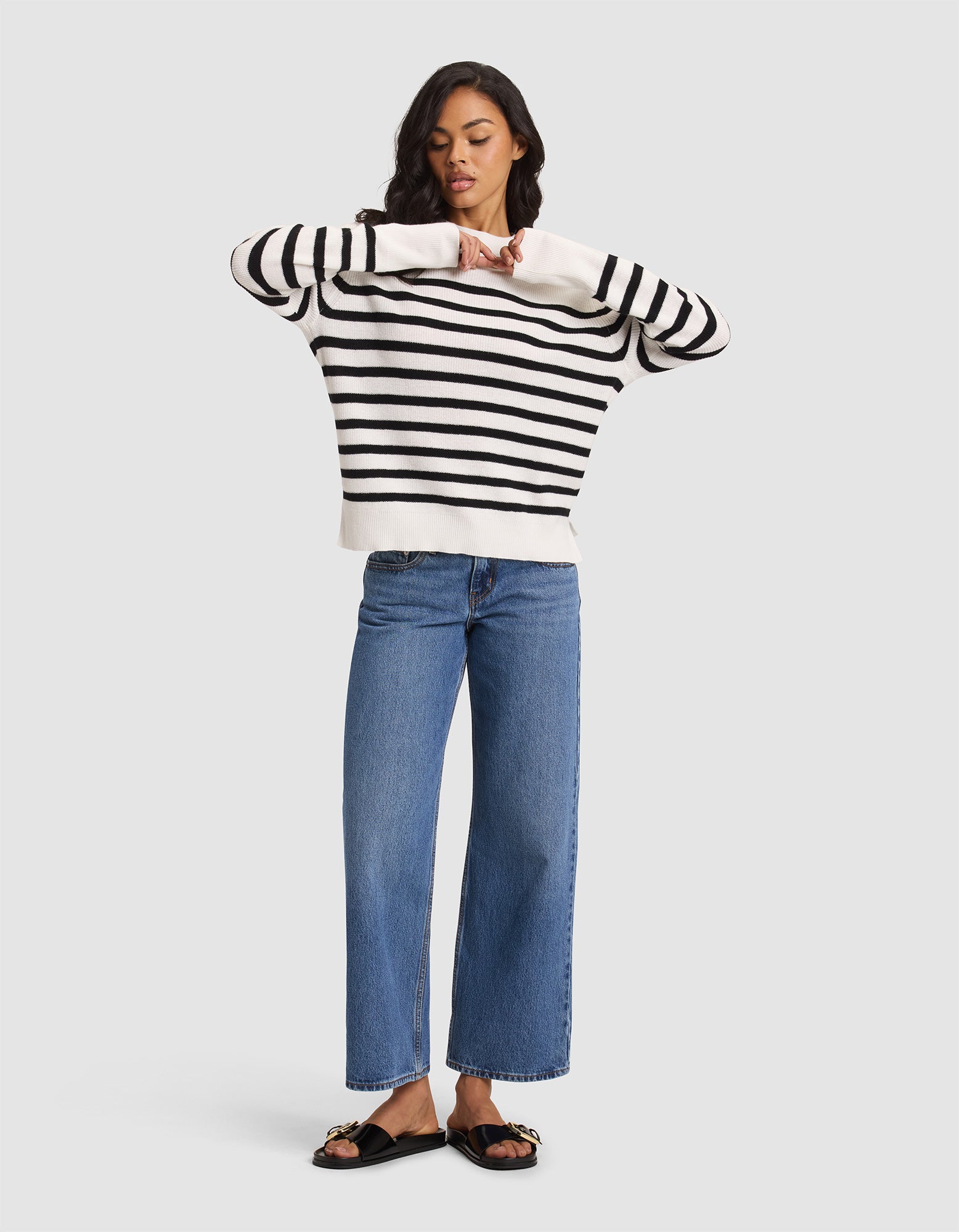 A woman with medium-length dark hair looks down, adjusting her sleeves as she models the Cozy Earth Women's Rowan Sweater against a plain background, styled with blue wide-leg jeans and black sandals featuring gold chain details. 