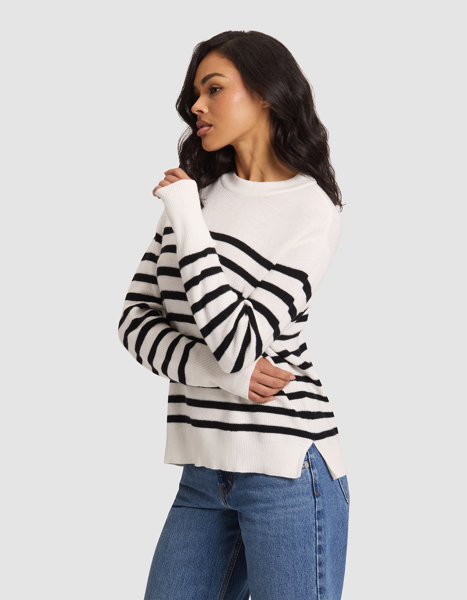 A woman with long dark hair models the Cozy Earth Women's Rowan Sweater, featuring black horizontal stripes on white, paired with blue jeans against a plain light gray background. She looks to the side, one hand touching her shoulder.