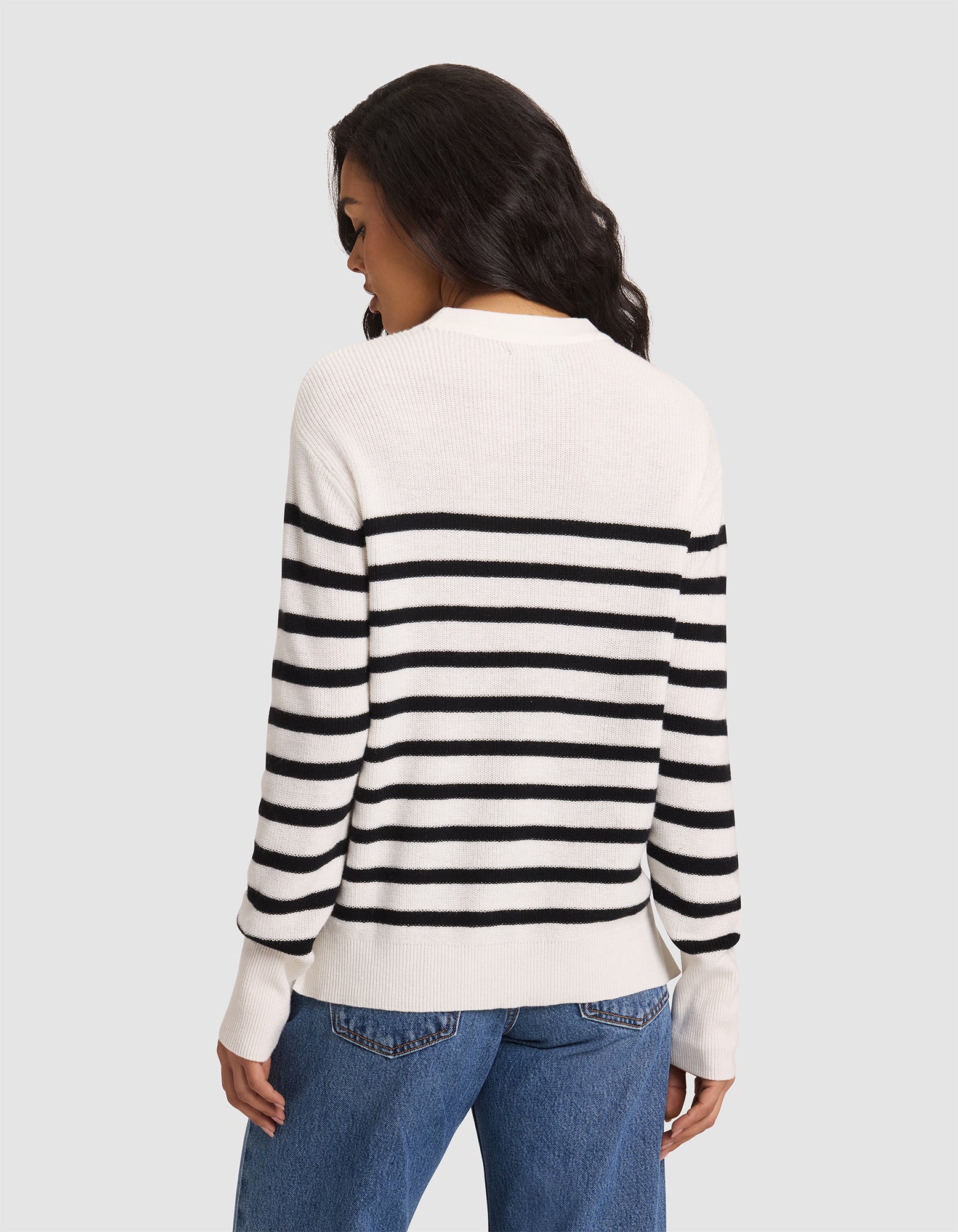 A woman with wavy dark hair, dressed in the Cozy Earth Women's Rowan Sweater—a white top with black horizontal stripes—and blue jeans stands facing away against a plain light gray background. 