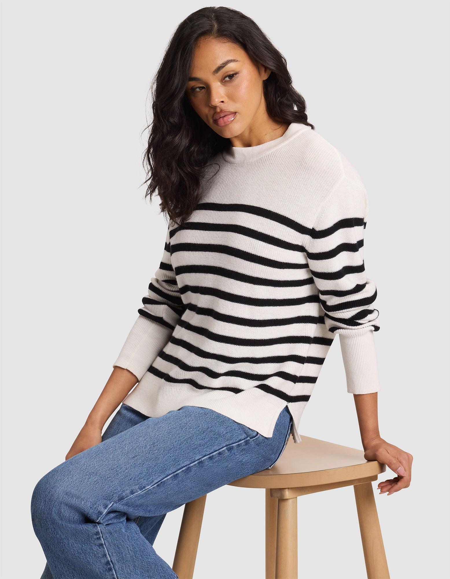 A woman with long dark hair sits on a wooden stool, wearing Cozy Earth's Women's Rowan Sweater in Bone/Jet Black Stripe and blue jeans, against a plain light gray background. 