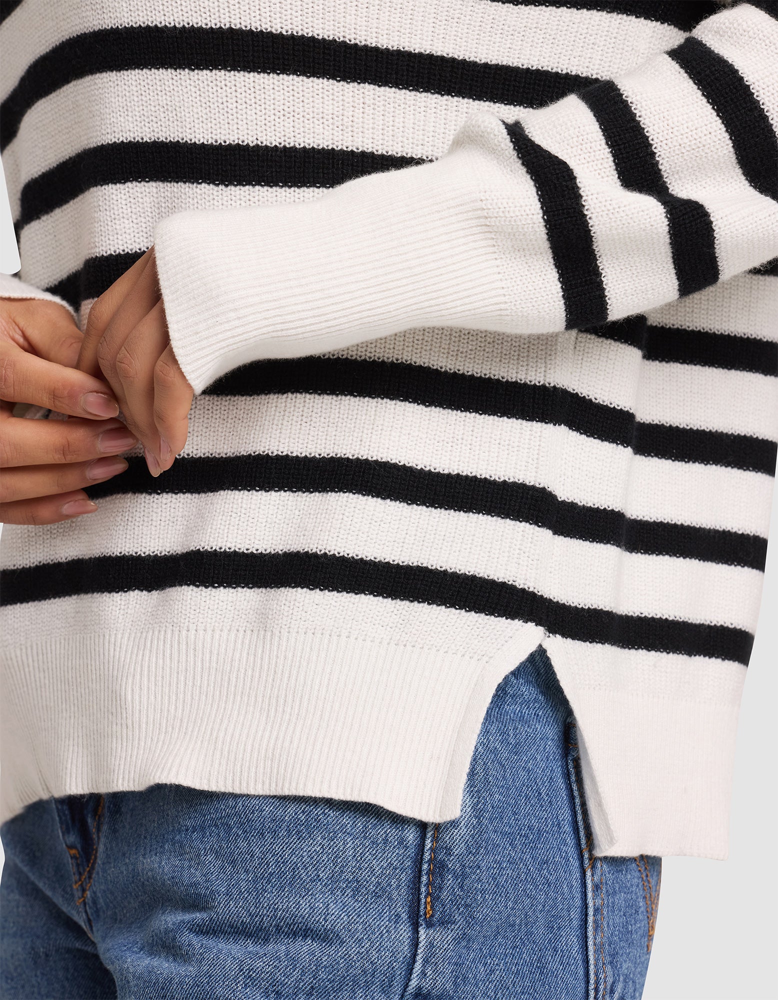 A woman wears the Cozy Earth Women's Rowan Sweater—white with black stripes and a side slit hem—paired with blue jeans, hands clasped near her waist. 