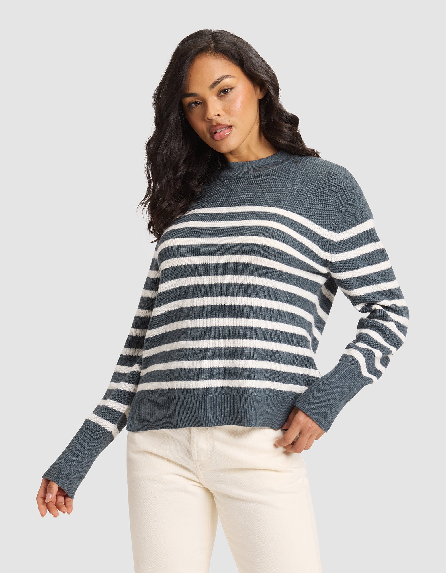 A woman with long dark hair models the Cozy Earth Women's Rowan Sweater—a blue and white striped knit—paired with off-white pants against a plain light gray background, one hand in her pocket and a relaxed expression. 