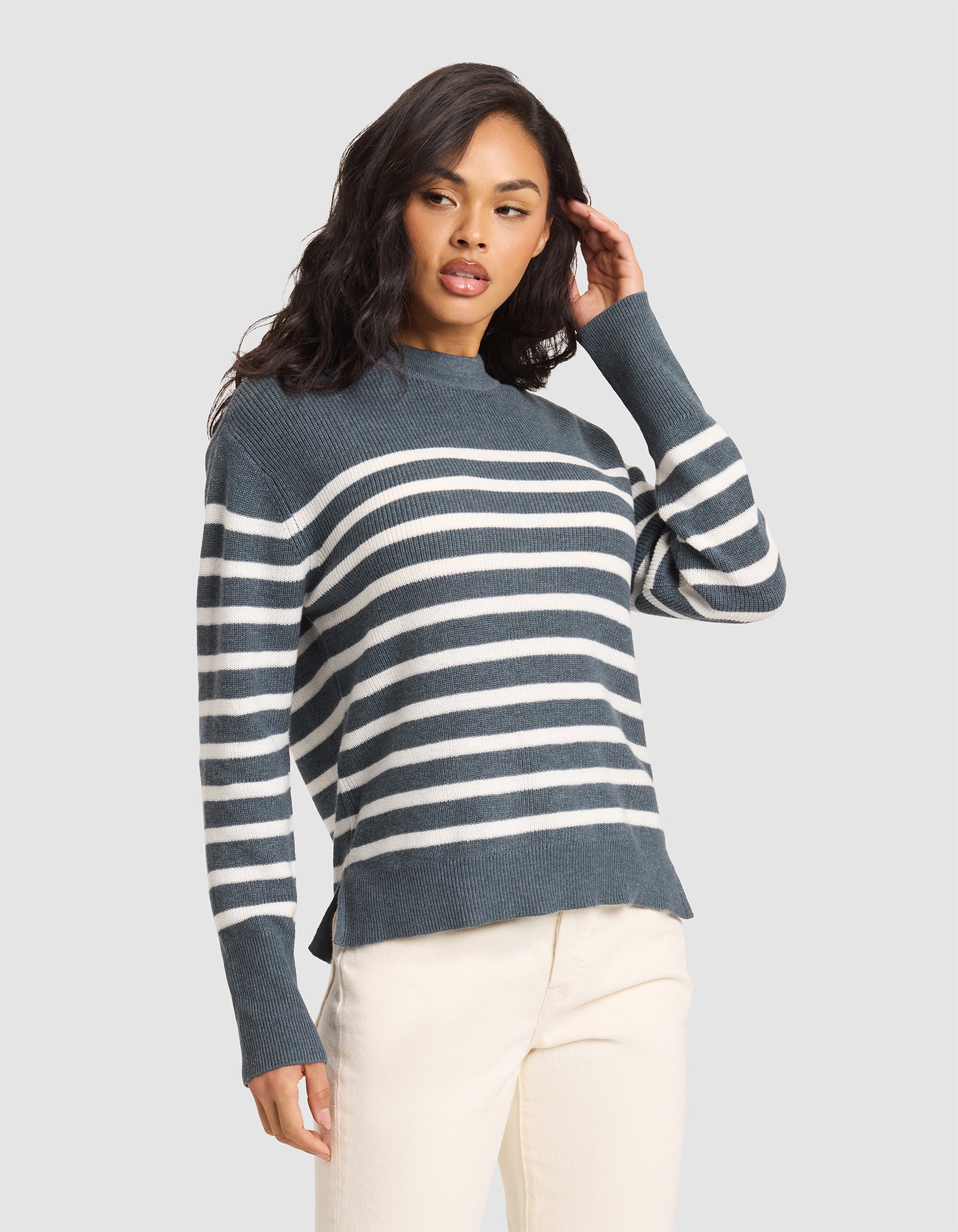 A woman poses with one hand in her hair against a plain light gray background, wearing the Cozy Earth Women's Rowan Sweater in Dawn/Bone Stripe and ivory pants. 