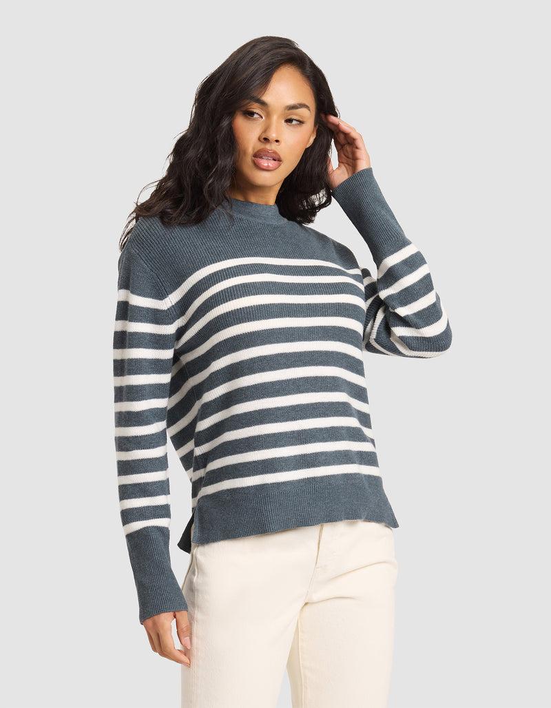 A woman poses with one hand in her hair against a plain light gray background, wearing the Cozy Earth Women's Rowan Sweater in Dawn/Bone Stripe and ivory pants.