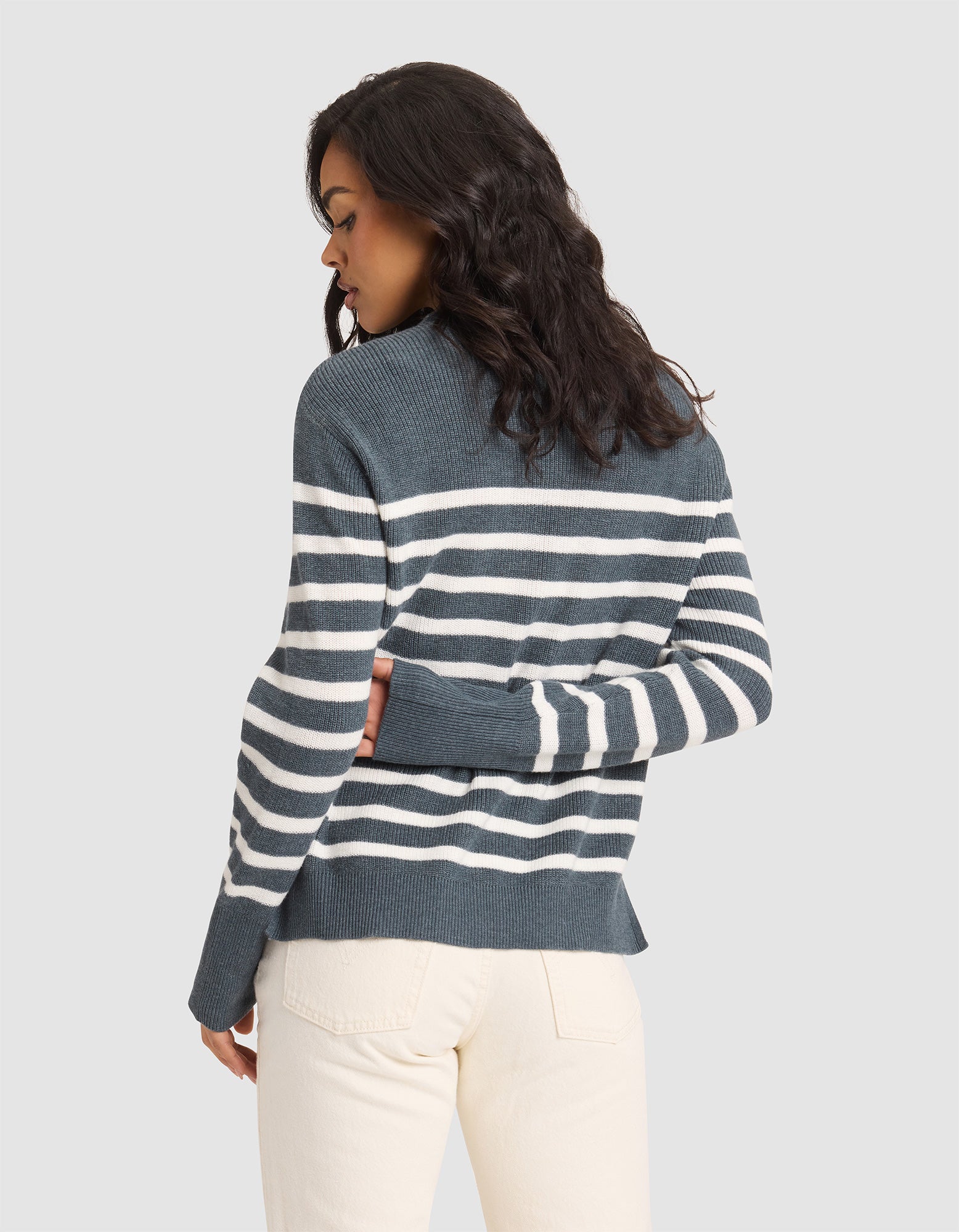 A woman with wavy dark hair models the Cozy Earth Women's Rowan Sweater, blue and white striped, paired with light beige pants. She stands with her back to the camera, head turned left, in front of a plain light background. 