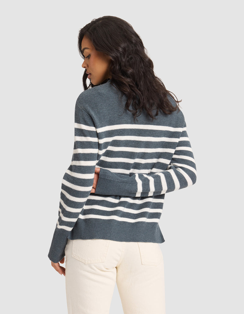 A woman with wavy dark hair models the Cozy Earth Women's Rowan Sweater, blue and white striped, paired with light beige pants. She stands with her back to the camera, head turned left, in front of a plain light background.