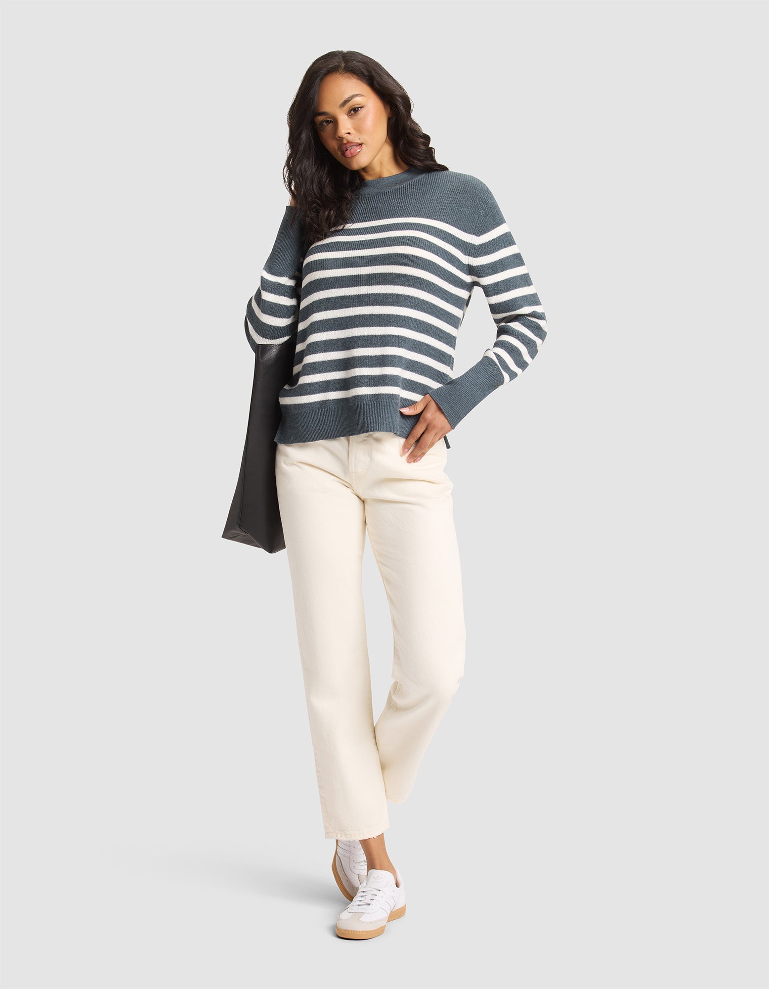 A woman poses in the Cozy Earth Women's Rowan Sweater, featuring dark green and white stripes. She pairs it with off-white pants and white sneakers, holding a dark jacket over her shoulder against a light gray background. 