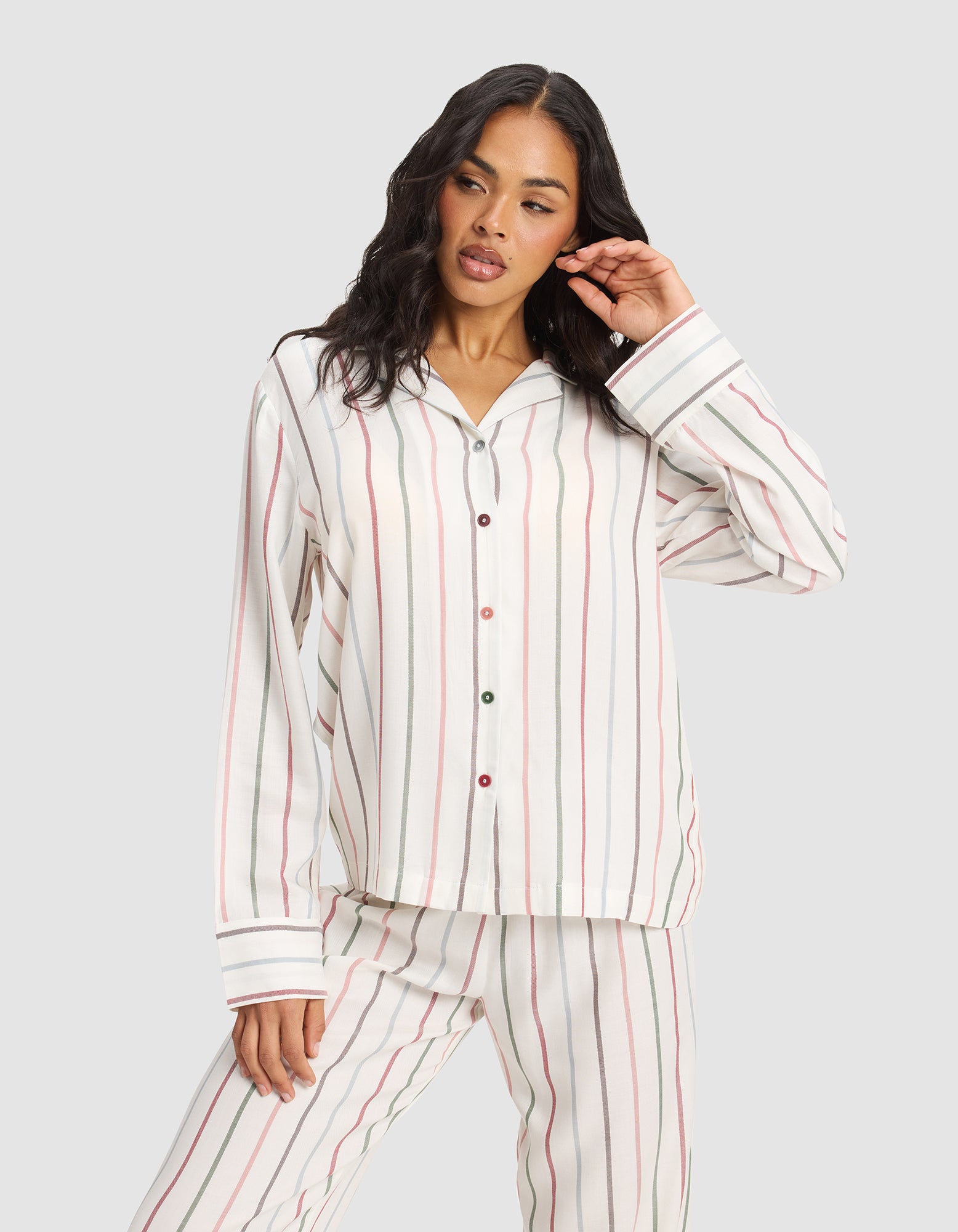 A woman wearing the Cozy Earth Women's Soft Woven Long Sleeve Pajama Top in white with vertical red, green, and beige stripes poses with one hand touching her hair against a plain light gray background. 