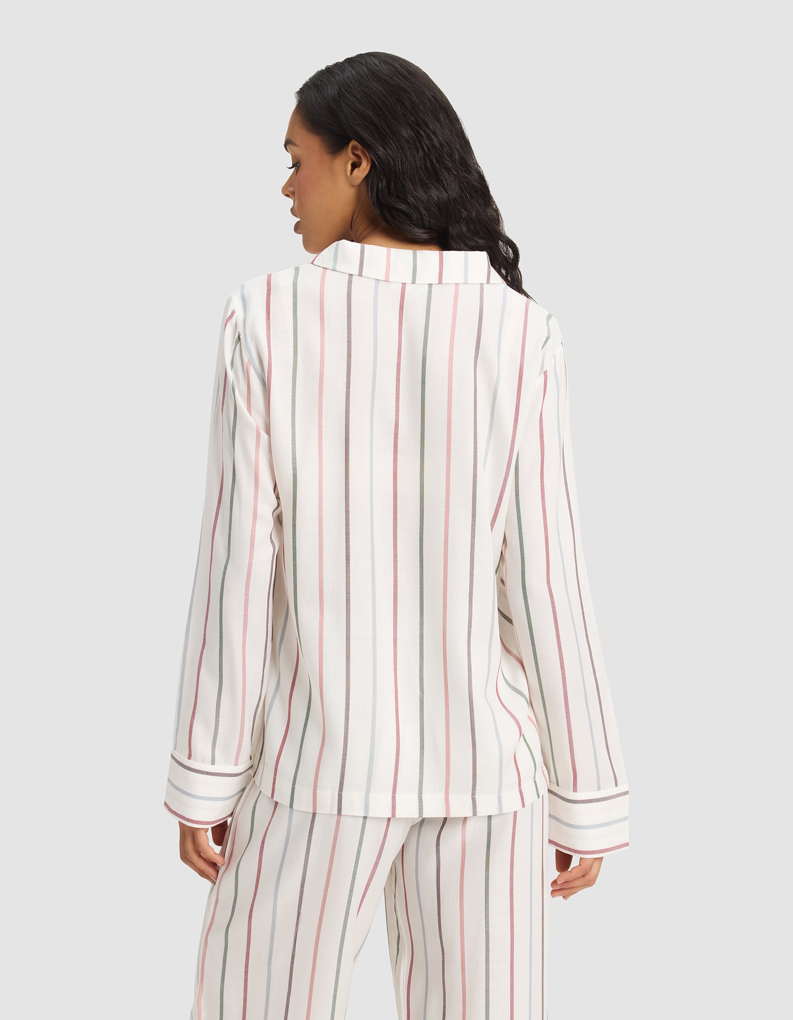 A woman with long dark hair faces away, modeling Cozy Earth's Women's Soft Woven Long Sleeve Pajama Top in a white fabric with vertical red, black, and beige stripes. The background is plain light gray. 