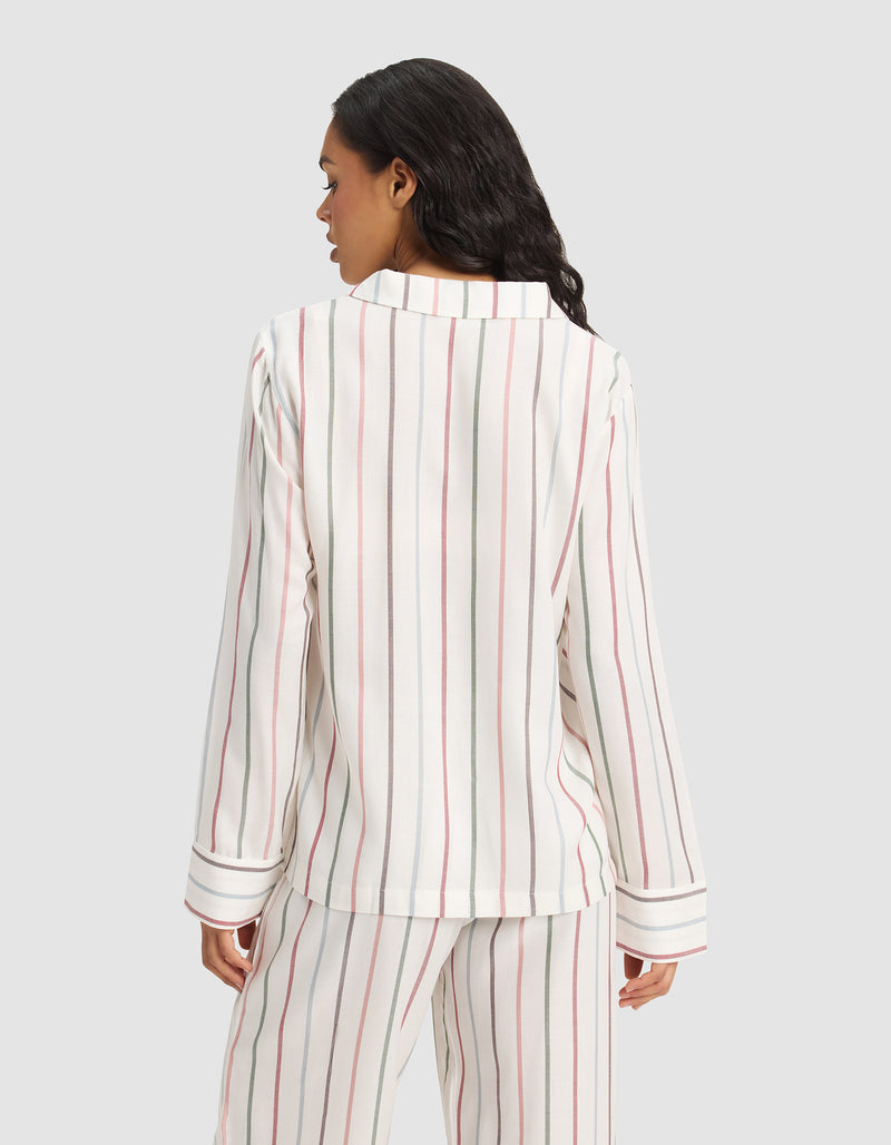 A woman with long dark hair faces away, modeling Cozy Earth's Women's Soft Woven Long Sleeve Pajama Top in a white fabric with vertical red, black, and beige stripes. The background is plain light gray.