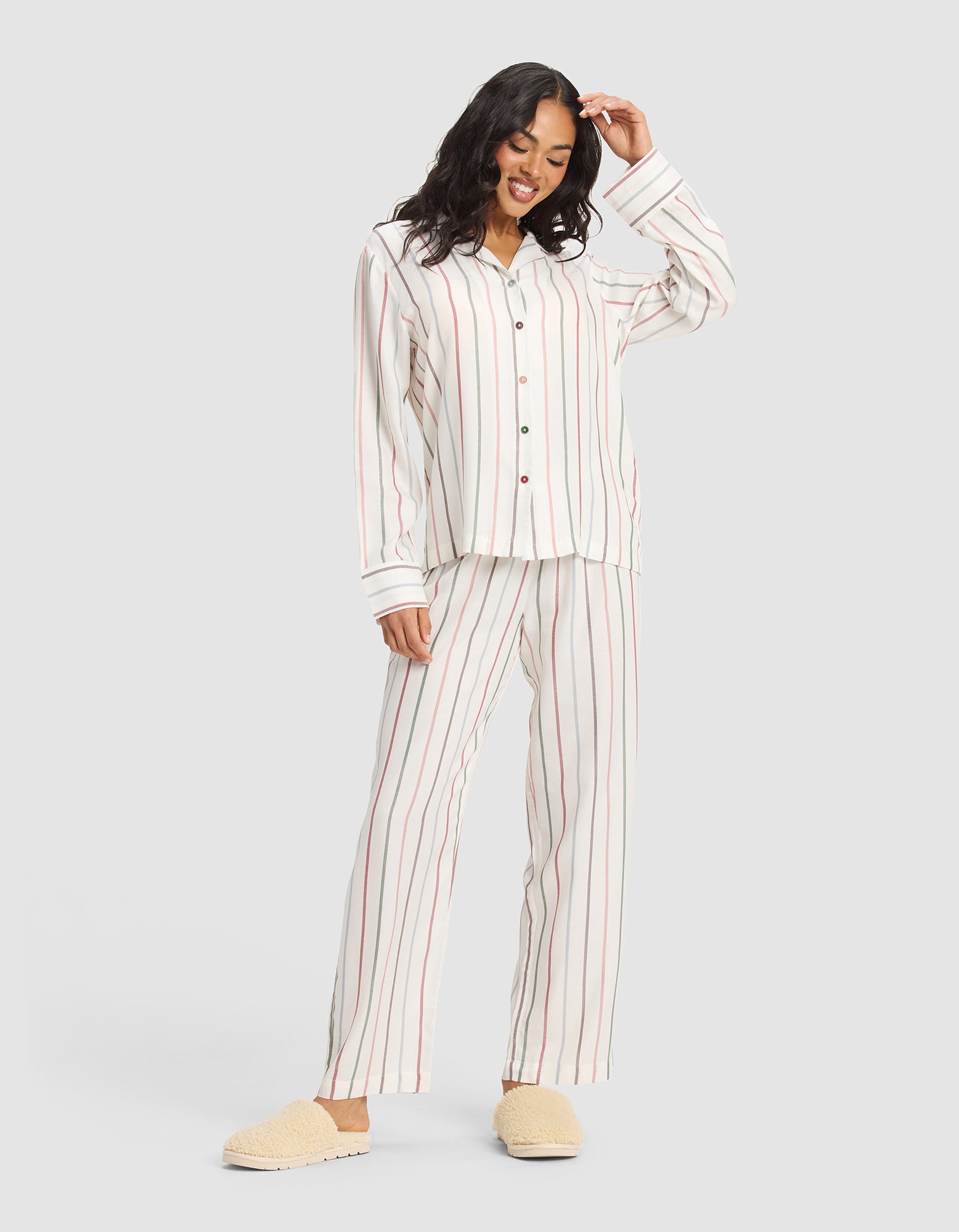 A woman smiles in a plain light gray setting, wearing the Cozy Earth Women's Soft Woven Long Sleeve Pajama Top in cream with vertical red and beige stripes, paired with beige slippers. 