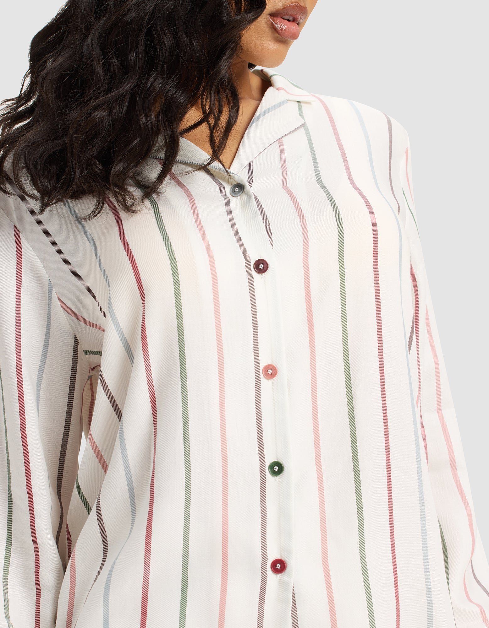 A woman models the Cozy Earth Women's Soft Woven Long Sleeve Pajama Set—a loose, white button-up top with pastel pink, green, and gray stripes and colorful buttons. Her wavy dark hair is visible; the image is cropped at her mouth and shoulders.