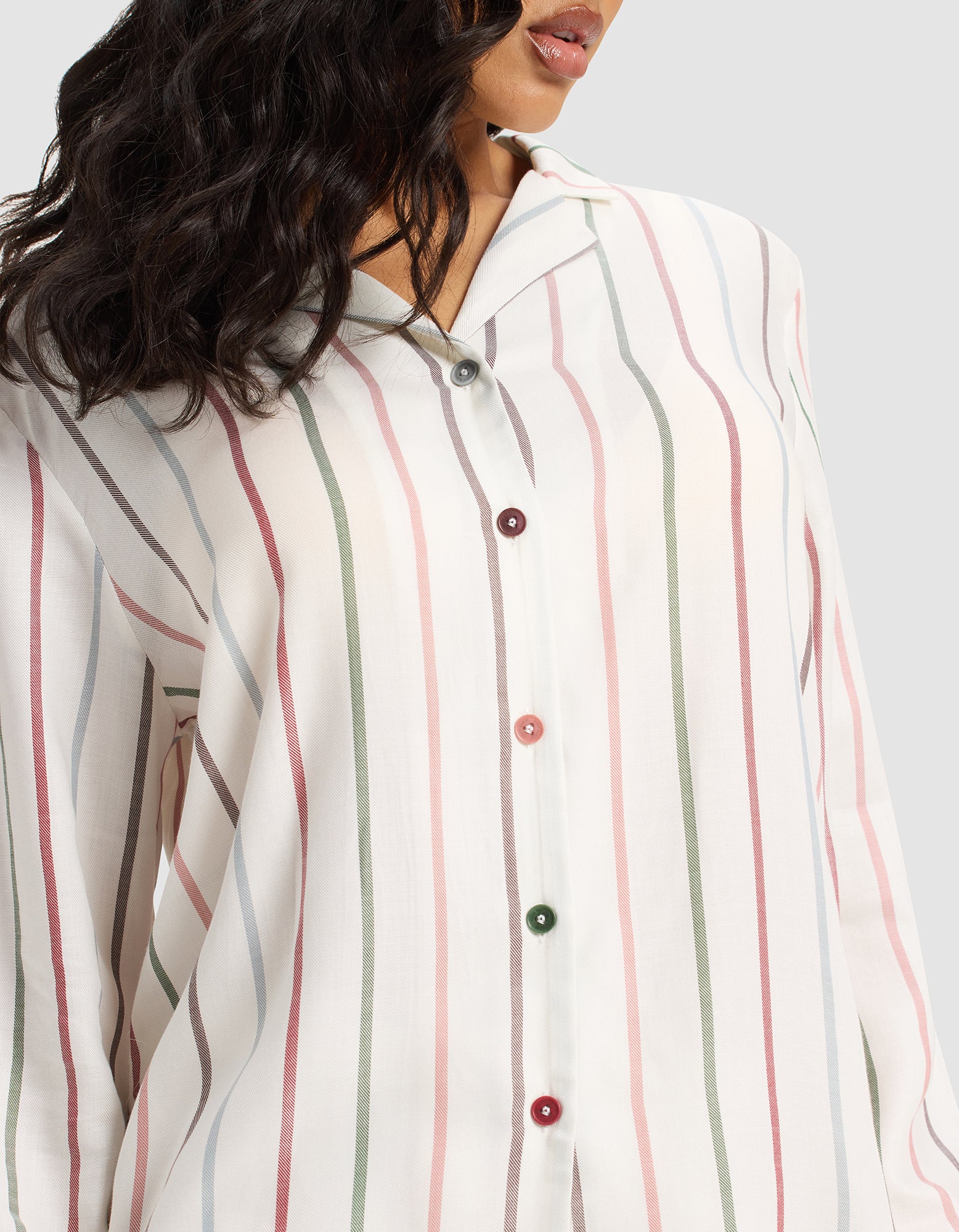 A woman models the Cozy Earth Women's Soft Woven Long Sleeve Pajama Set—a loose, white button-up top with pastel pink, green, and gray stripes and colorful buttons. Her wavy dark hair is visible; the image is cropped at her mouth and shoulders.