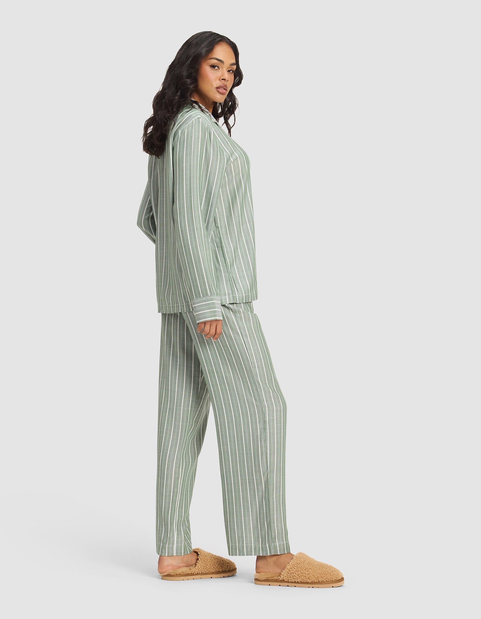 A woman with long, wavy dark hair models the Cozy Earth Women's Soft Woven Long Sleeve Pajama Set in green and white stripes with matching pants and shirt, paired with tan textured slippers, standing calmly against a plain background.