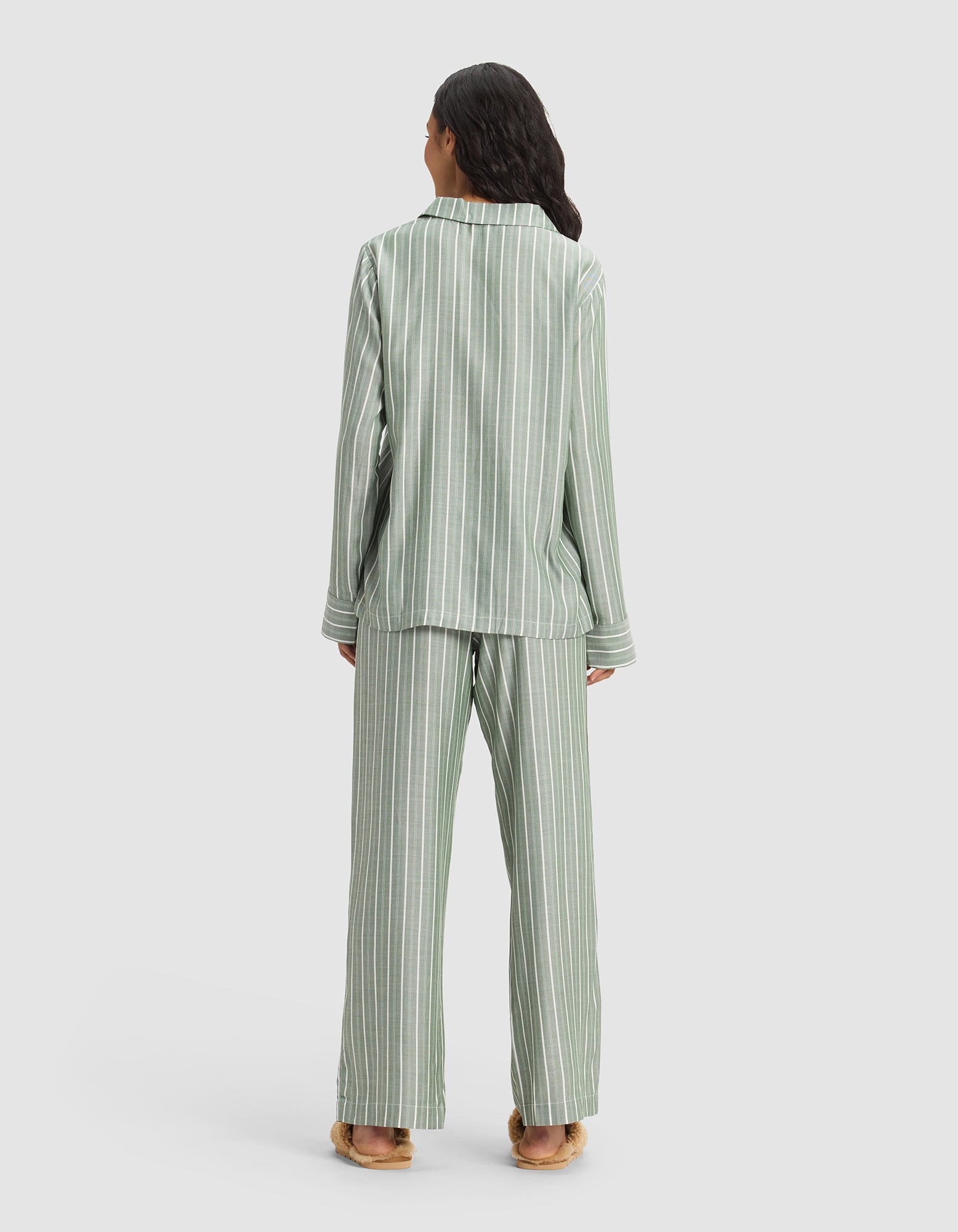 A person stands facing away, wearing the Cozy Earth Women's Soft Woven Long Sleeve Pajama Set in light green with white stripes and tan slippers, set against a plain light background.