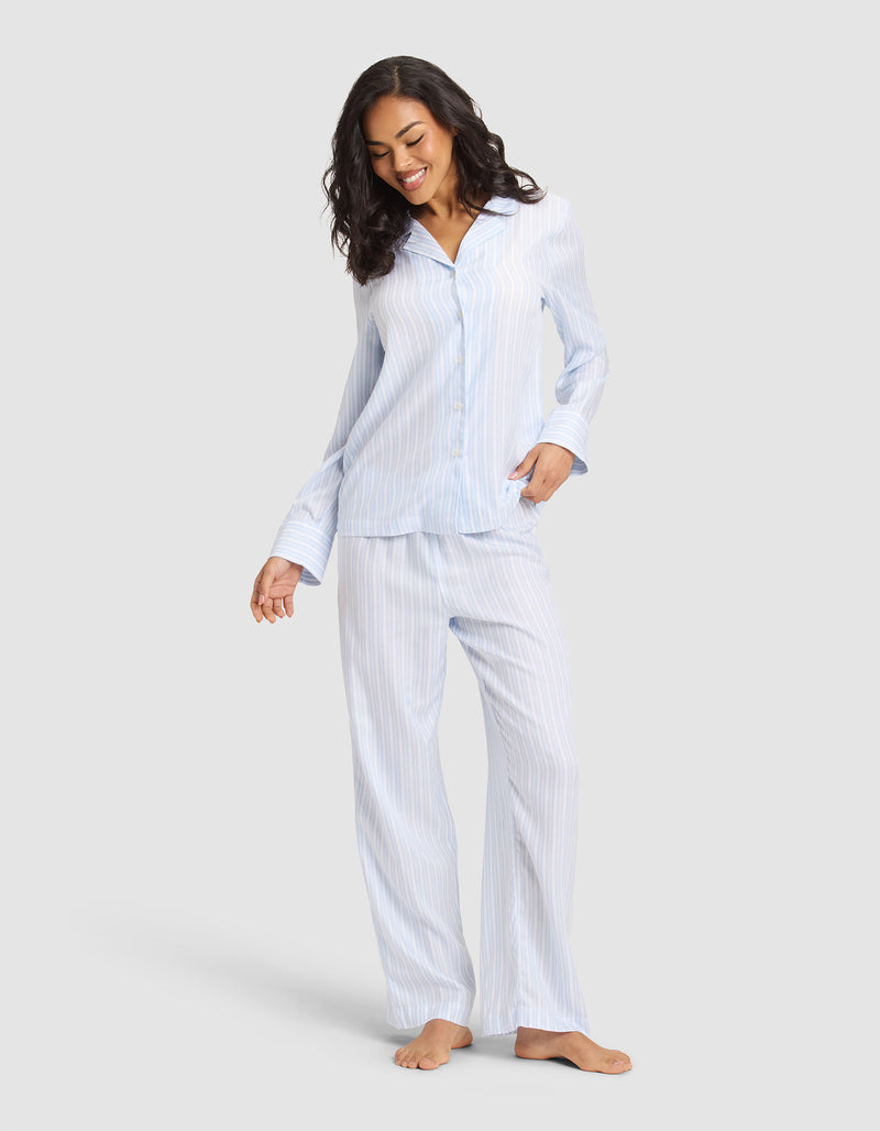 A woman smiles and looks down, barefoot in the Cozy Earth Women's Soft Woven Long Sleeve Pajama Set—light blue and white striped with a button-up shirt and long pants.