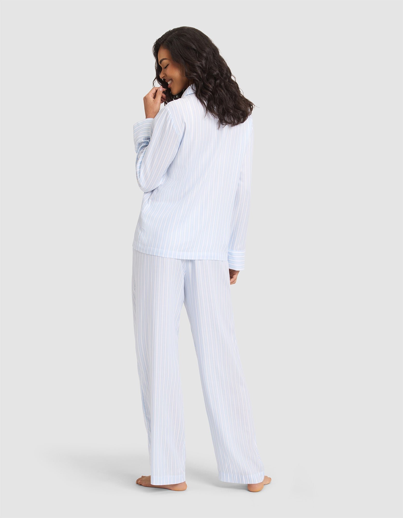 A woman stands barefoot, facing away in the Cozy Earth Women's Soft Woven Long Sleeve Pajama Set, light blue and white striped. She glances over her shoulder with a smile, hand near her mouth, against a plain light gray background. 