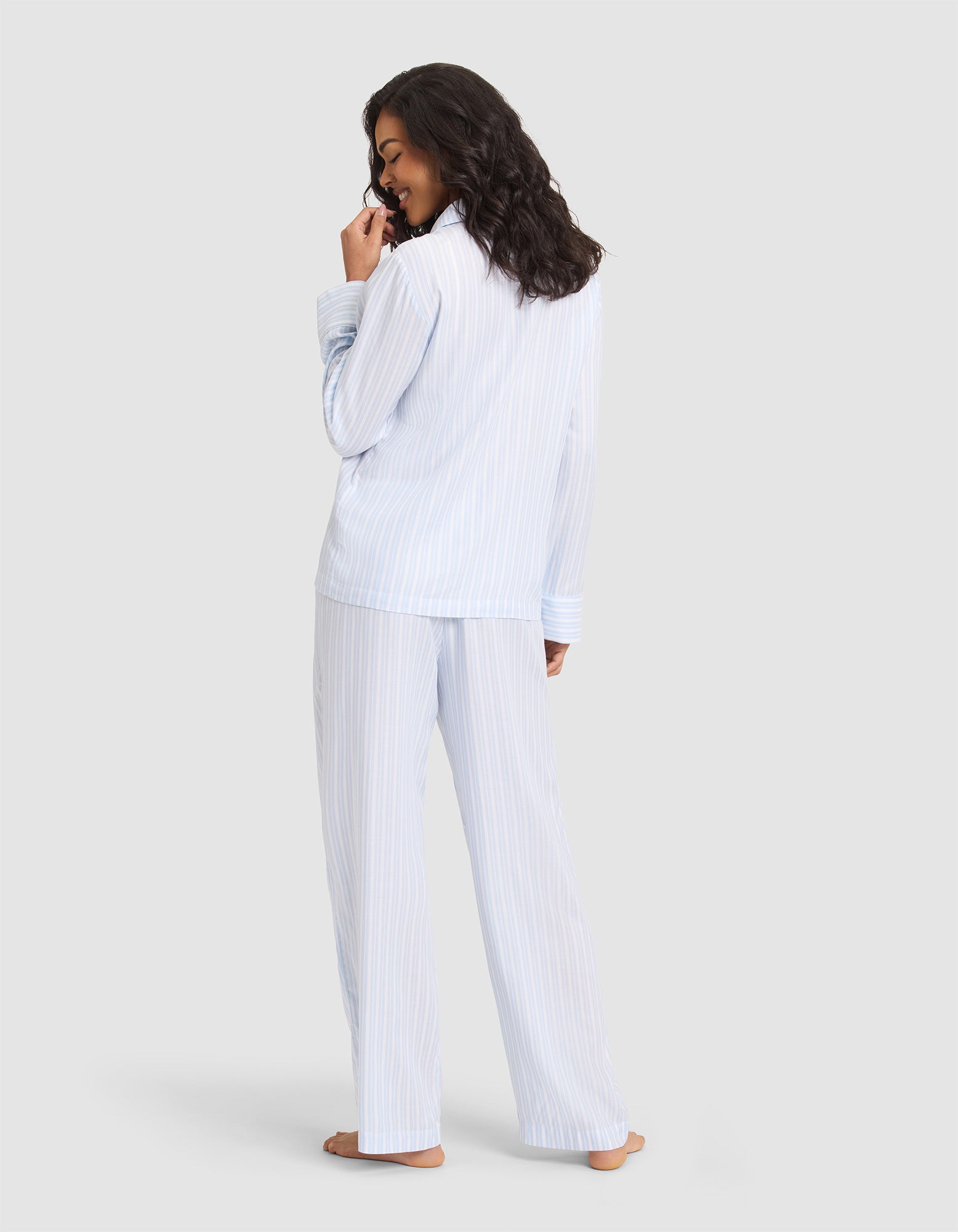 A woman stands barefoot, facing away in the Cozy Earth Women's Soft Woven Long Sleeve Pajama Set, light blue and white striped. She glances over her shoulder with a smile, hand near her mouth, against a plain light gray background. 