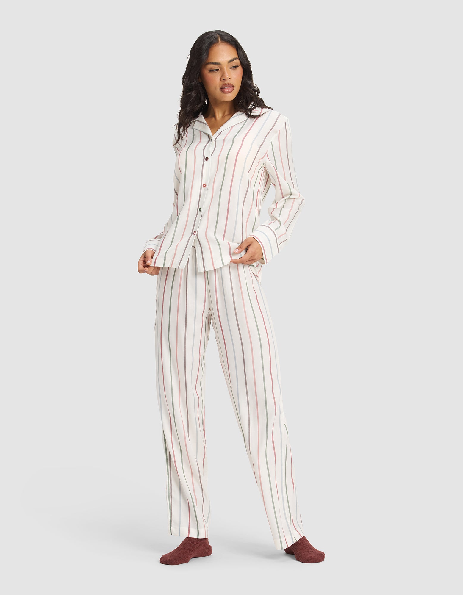 A woman wears the Cozy Earth Women's Soft Woven Long Sleeve Pajama Set, featuring white fabric with red and beige vertical stripes, a button-up collared top, and paired with maroon socks for a comfortable, relaxed look.
