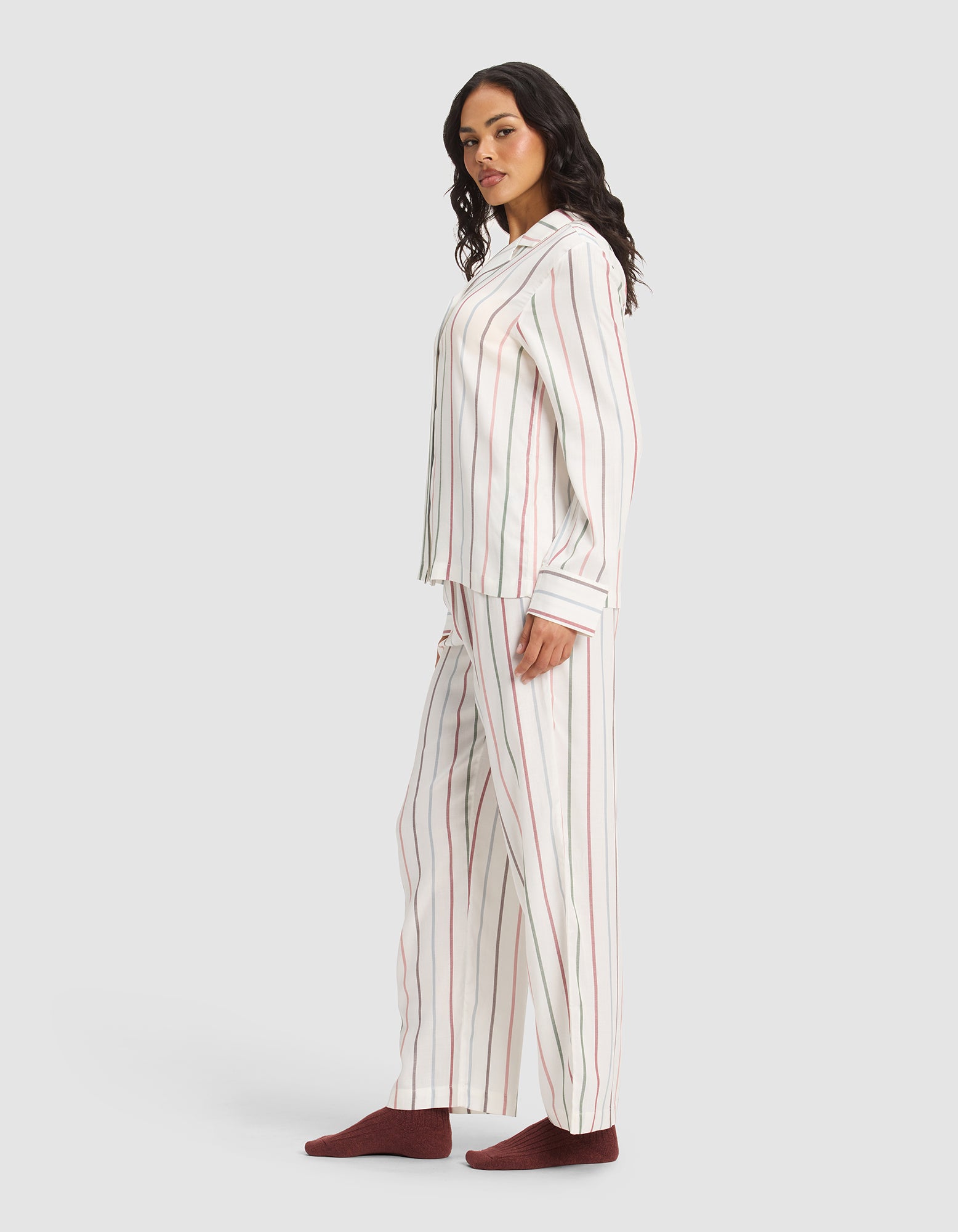 A woman stands sideways wearing the Cozy Earth Women's Soft Woven Long Sleeve Pajama Set with vertical red and gray stripes, paired with dark red socks, against a plain light gray background.